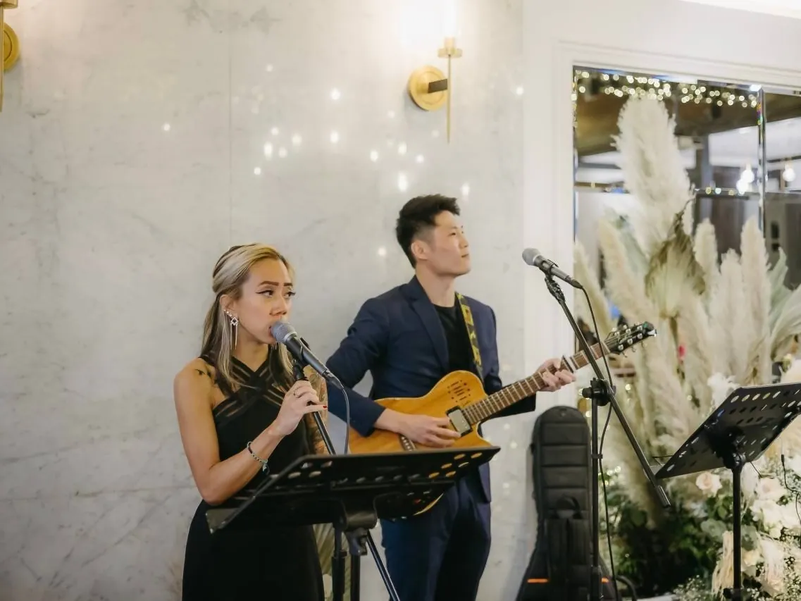 NyaLi singing and Ze on acoustic guitar at a wedding, a marble wall and pampas grass behind