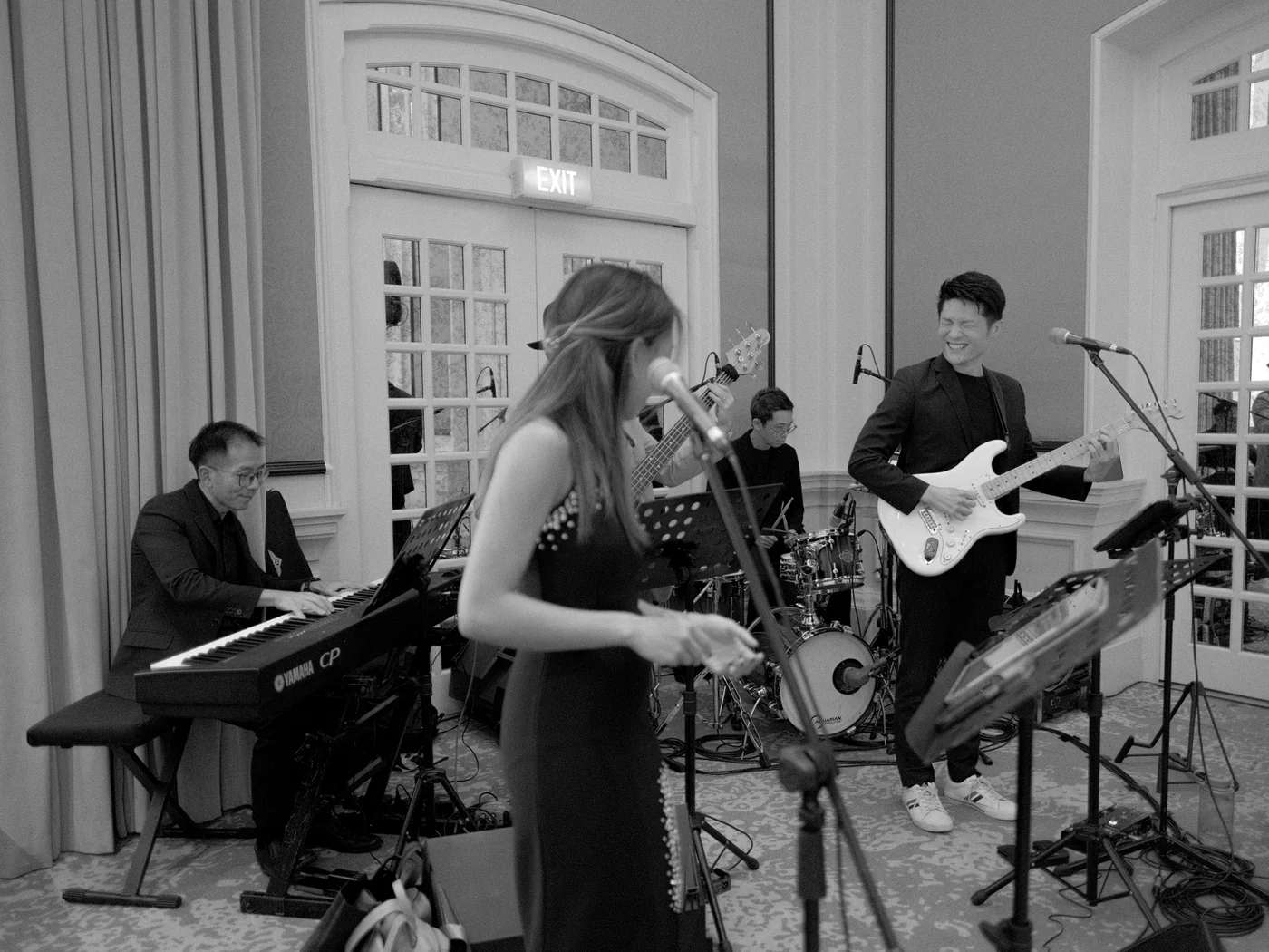 The full band at an indoor event, in black and white: NyaLi singing, Ze laughing on guitar, with keys, drums and bass behind